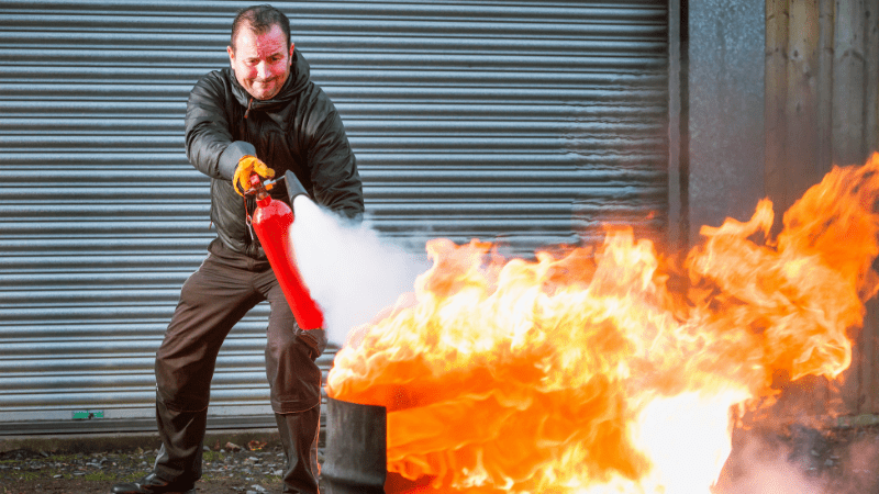 Na foto, um homem usando o extintor de incêndio para amenizar a propagação do fogo, sendo uma das dicas da prevenção de incêndios.